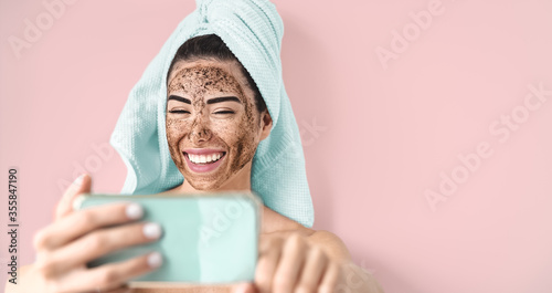 Young smiling woman taking selfie while doing coffee scrub facial mask - Happy girl having skin care spa day at home - Healthy alternative natural exfoliation treatment and technology concept photo