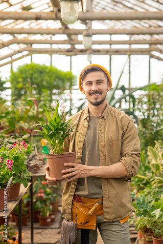 Portrait of cheerful young greenhouse man with beard and mustache holding potted plant in hothouse