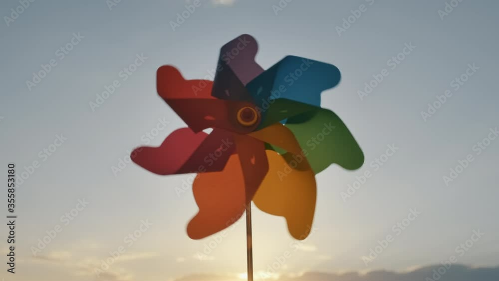 Pinwheel rotating colored plastic with blowing wind against a blue sky, white clouds, silhouettes of city buildings on sunset slow motion. Symbol of freedom and life. Toy spinner. Bright disk of sun Pinwheel rotating colored plastic with blowing wind against a blue sky, white clouds, silhouettes of city buildings on sunset slow motion. Symbol of freedom and life. Toy spinner. Bright disk of sun