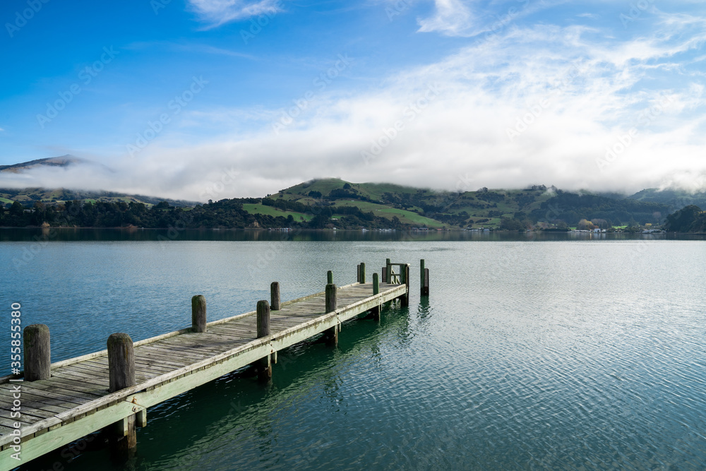 Wooden pier and sailboat at Akaroa harbor, New Zealand. Clear sky with a few white clouds and mist. Symbol for relaxation, wealth, leisure activity. Panorama format.