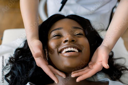 Close-up of hands of cosmetologist woman making special face massage to young pretty African smiling woman lying on couch in beauty salon. Concept of antiaging facial skin treatments and massage photo