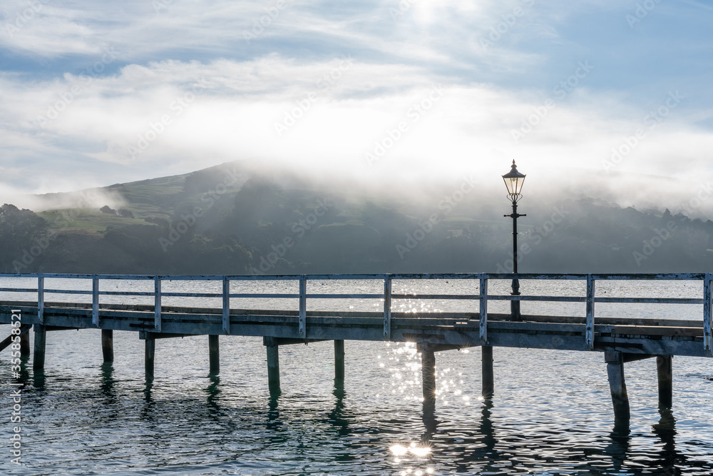 Fototapeta premium Wooden pier and sailboat at Akaroa harbor, New Zealand. Clear sky with a few white clouds and mist. Symbol for relaxation, wealth, leisure activity. Panorama format.