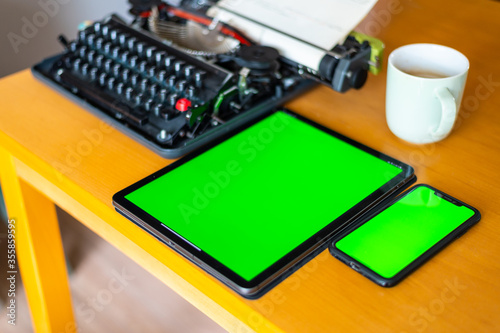 next to an old typewriter lies a tablet with a green screen on the table, a cell phone and a coffee