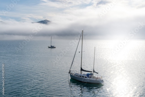 Fototapeta Naklejka Na Ścianę i Meble -  Blue sky, white clouds, mist & aquamarine seas at Akaroa Harbor. Anchored are fishing boats & sailboats, along the Port at Akaroa, Canterbury, New Zealand.