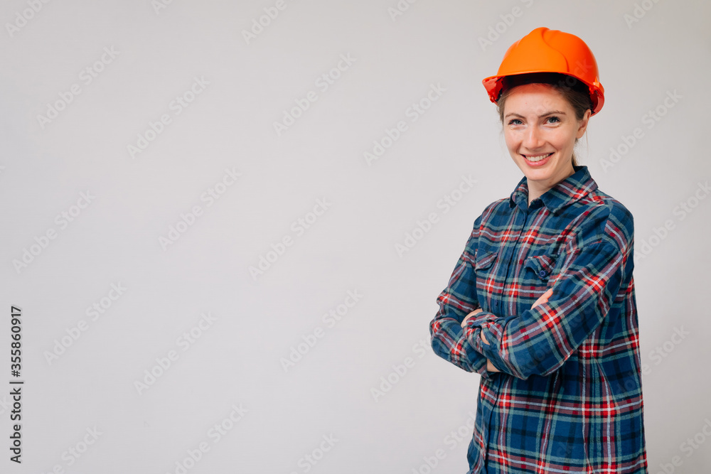 Portrait of a young smiling girl builder in a plaid shirt in an orange protective helmet against a light background Portrait of a young smiling girl builder in a plaid shirt in an orange protective helmet against a light background