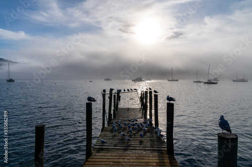 Wooden pier and sailboat at Akaroa harbor, New Zealand. Clear sky with a few white clouds and mist. Symbol for relaxation, wealth, leisure activity. Panorama format.