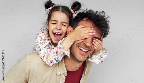 Indoor image of playful daughter covers the eyes with hands of her father for a surprise on Father's Day. Handsome man playing with kid peekaboo game, shares love together. photo