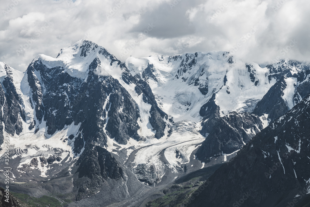 Atmospheric alpine landscape with massive hanging glacier on giant mountain. Big glacier tongue on mountainside. Low clouds among snowbound mountains. Cracks on ice. Majestic scenery on high altitude.