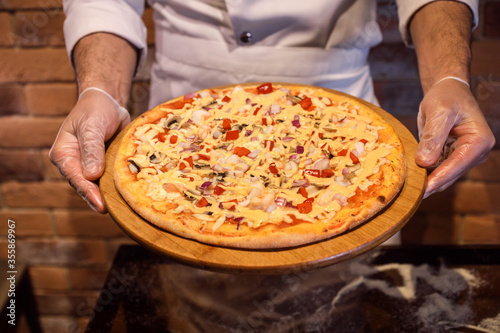 Chef in gloves holds appetizing pizza on the wooden stand, tablet. Pizza is made of shrimps, onion, paprica, cheese sauce. 