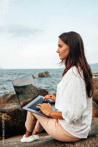 Young successful, smiling girl working using a laptop, sitting on a rocky seashore. traveling, blogger, freelancer, content plan, work online