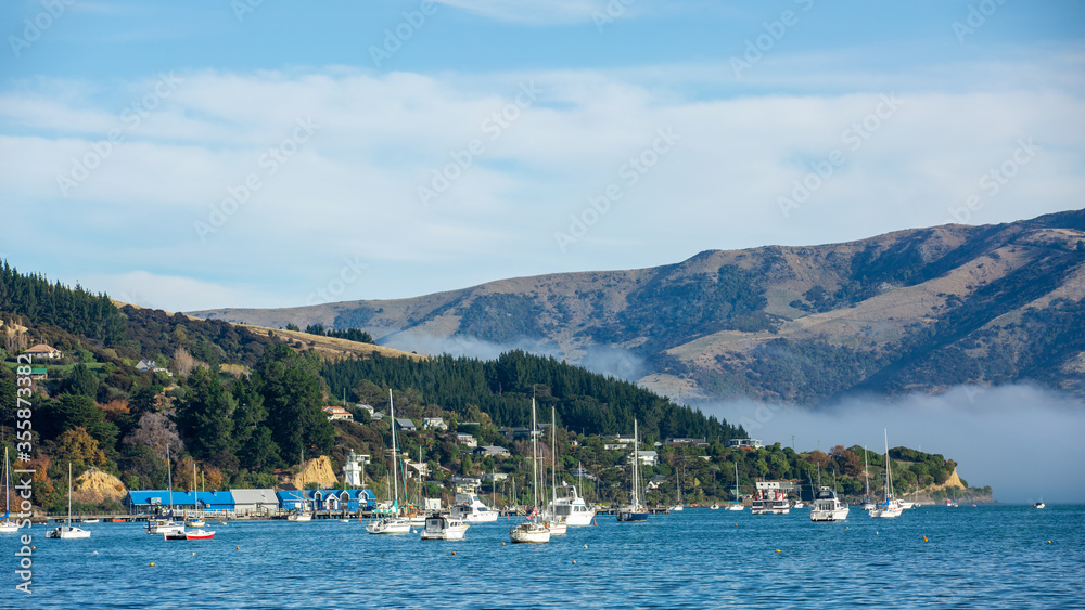 Blue sky, white clouds, mist & aquamarine seas at Akaroa Harbor. Anchored are fishing boats & sailboats, along the Port at Akaroa, Canterbury, New Zealand.