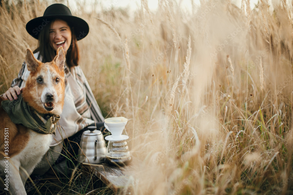 Φωτογραφία Stylish hipster traveler hugging her golden dog and smiling, brewing fresh coffee in rural countryside herbs in sunset light