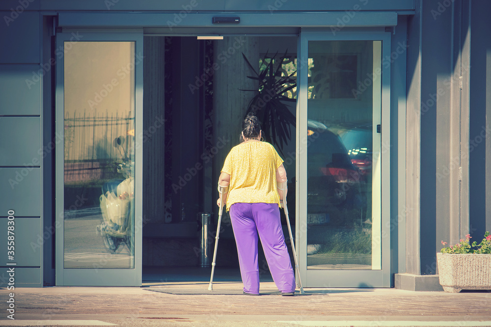 woman standing with crutches. She's looking the entrance door of a clinic
