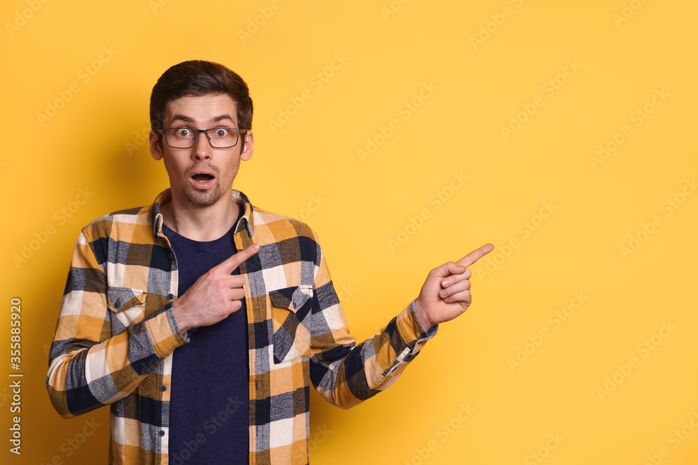 Shocked young man in eyeglasses keeps mouth opened, surprised, shows something, pointing with index fingers on blank copy space for advertising, isolated over yellow background. Advertisement concept Shocked young man in eyeglasses keeps mouth opened, surprised, shows something, pointing with index fingers on blank copy space for advertising, isolated over yellow background. Advertisement concept