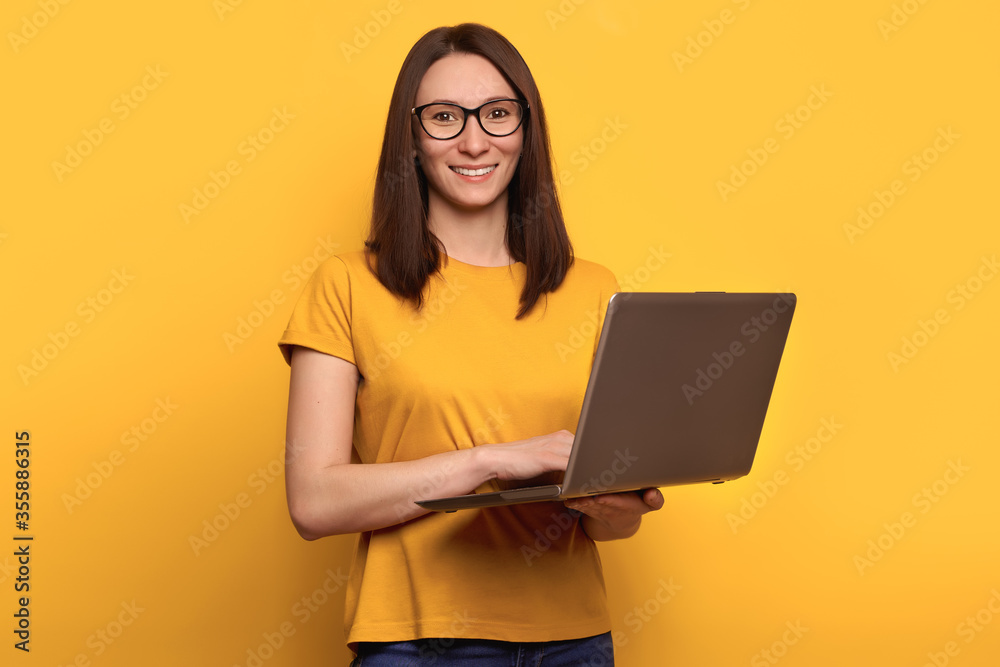 Young cute woman in eyeglasses is smiling happily, holding laptop isolated over yellow background, looking at the camera. Copy space. Promotion concept Young cute woman in eyeglasses is smiling happily, holding laptop isolated over yellow background, looking at the camera. Copy space. Promotion concept
