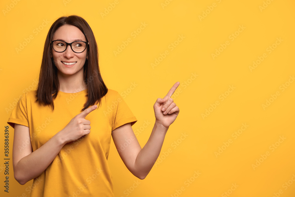 Happy young mixed race woman in glasses giving direction for advertising with index fingers, isolated on yellow background. Brunette female pointing at free space. Advert concept Happy young mixed race woman in glasses giving direction for advertising with index fingers, isolated on yellow background. Brunette female pointing at free space. Advert concept