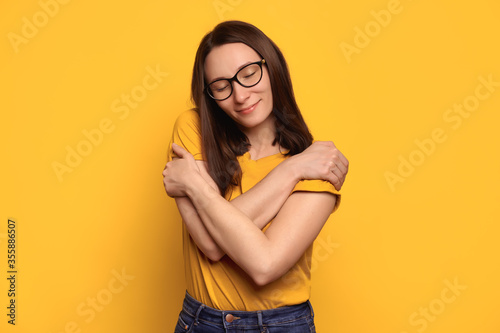 Young beautiful brunette woman in eyeglasses wearing casual yellow t-shirt hugging herself, looks calm and positive, smiling happily, love her body. Body positive, self acceptance, self esteem concept
