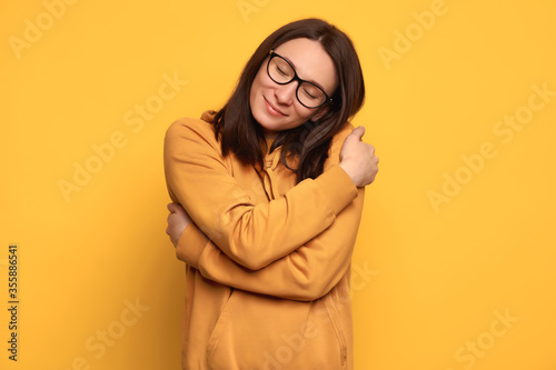 Closeup portrait confident smiling woman in glasses and orange cozy jumper holding and hugging herself isolated on yellow background. Positive human emotion. Love yourself and body positive concept photo