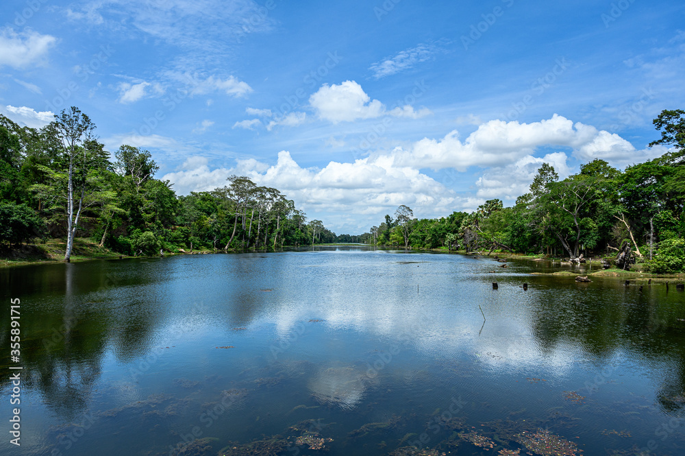 Fototapeta premium Scenic view of the river around the Angkor Thom temple in Siem Reap, Cambodia. River surrounded by trees, beautiful clean sky with clouds, reflection in the water, sunny day.