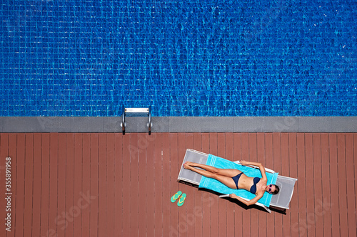 Enjoying suntan. Vacation concept. Top view of slim young woman in bikini on the sun lounger near the big swimming pool.
