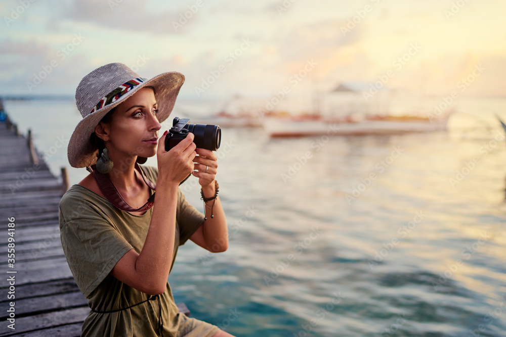 Obraz premium Photography and travel. Young woman in hat holding camera sitting on wooden fishing pier with beautiful tropical sea view.