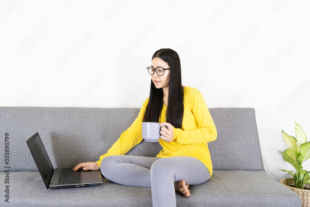 young asian woman in glasses using laptop, sitting on gray sofa in living room at home,female chatting in social network, shopping or working online, looking at computer screen,relax with coffee young asian woman in glasses using laptop, sitting on gray sofa in living room at home,female chatting in social network, shopping or working online, looking at computer screen,relax with coffee
