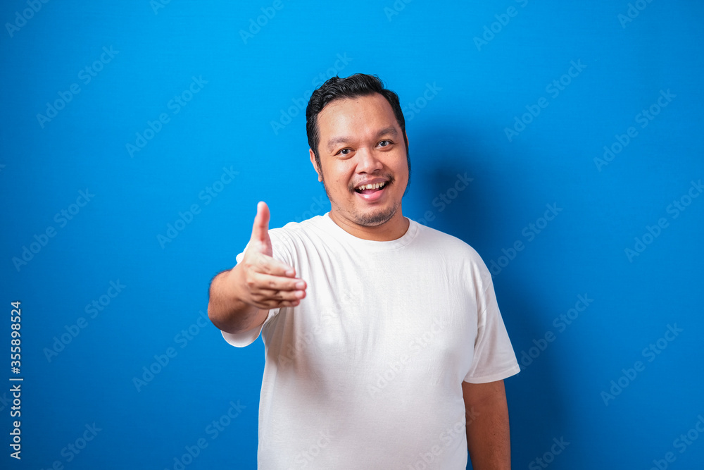 Portrait of young Asian man offering handshake and smiling against blue background, selective focus image, deal agreement concept