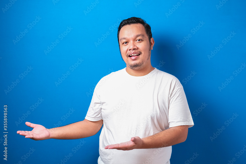 Portrait of funny young Asian man in white tshirt smiling and pointing to presenting something on his side, against blue background with copy space Portrait of funny young Asian man in white tshirt smiling and pointing to presenting something on his side, against blue background with copy space