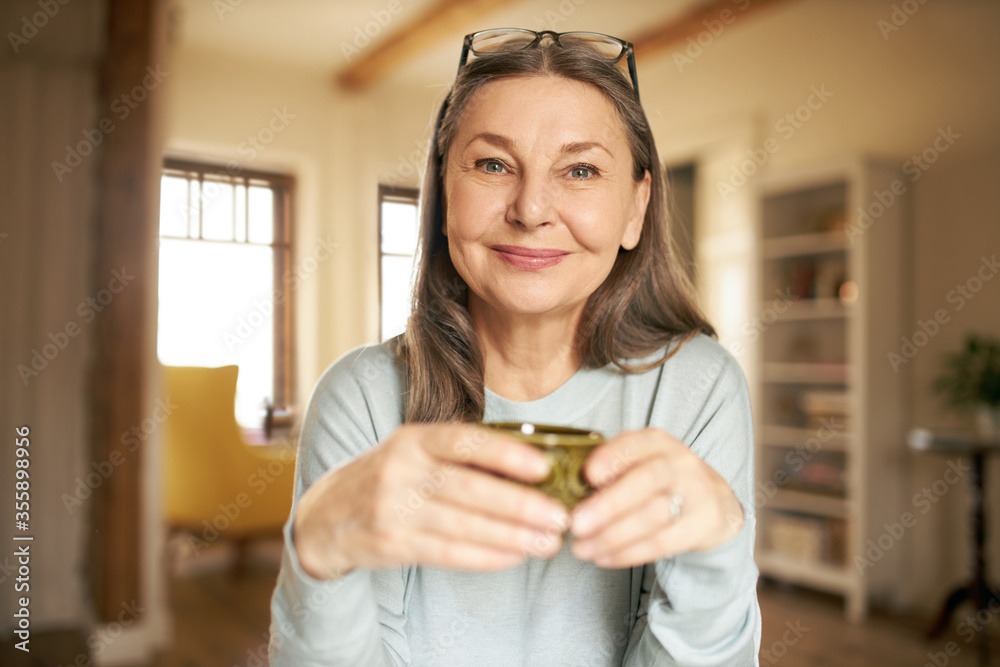 Φωτογραφία Indoor image of joyful beautiful senior woman with glasses on head sitting at home with cup in her hands, relaxing, drinking morning coffee, smiling