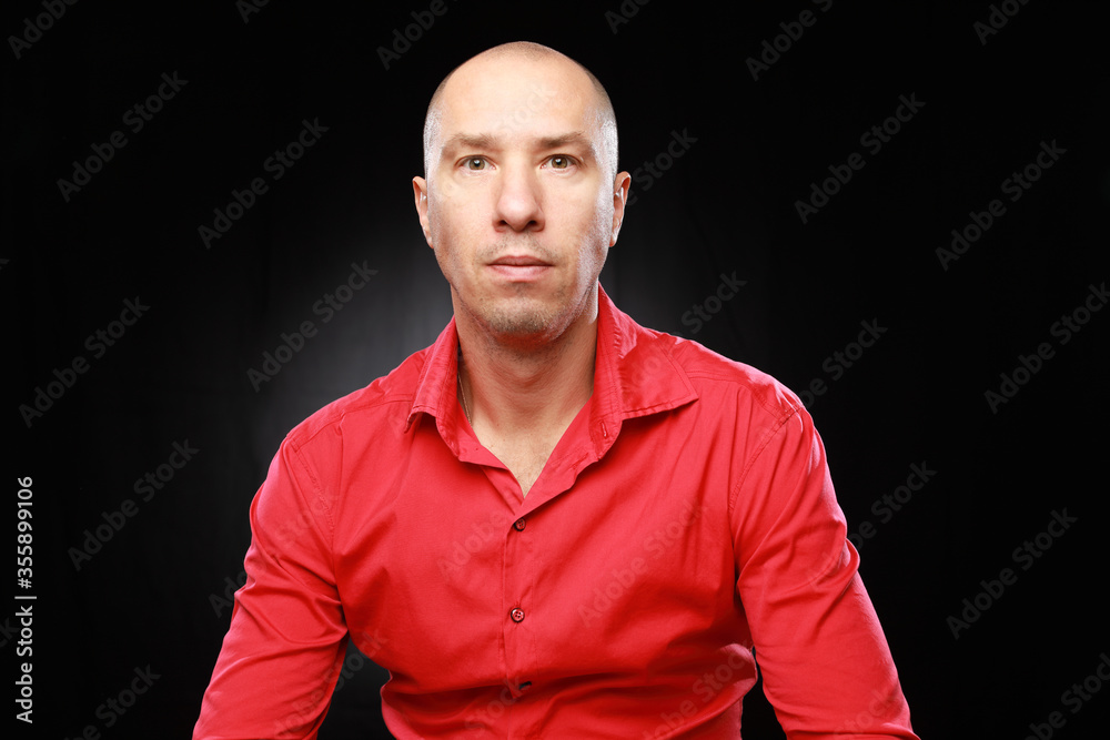 Headshot of bald adult man in red shirt with funny emotions isolated on black background with copy space.