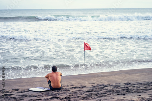 No swimming! Surfer man sitting next to the sea at the beach with red flag. photo