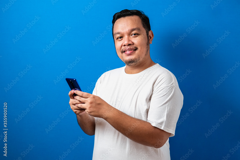 Portrait of Fat Asian man browsing Internet on smartphone, against blue background studio shot Portrait of Fat Asian man browsing Internet on smartphone, against blue background studio shot