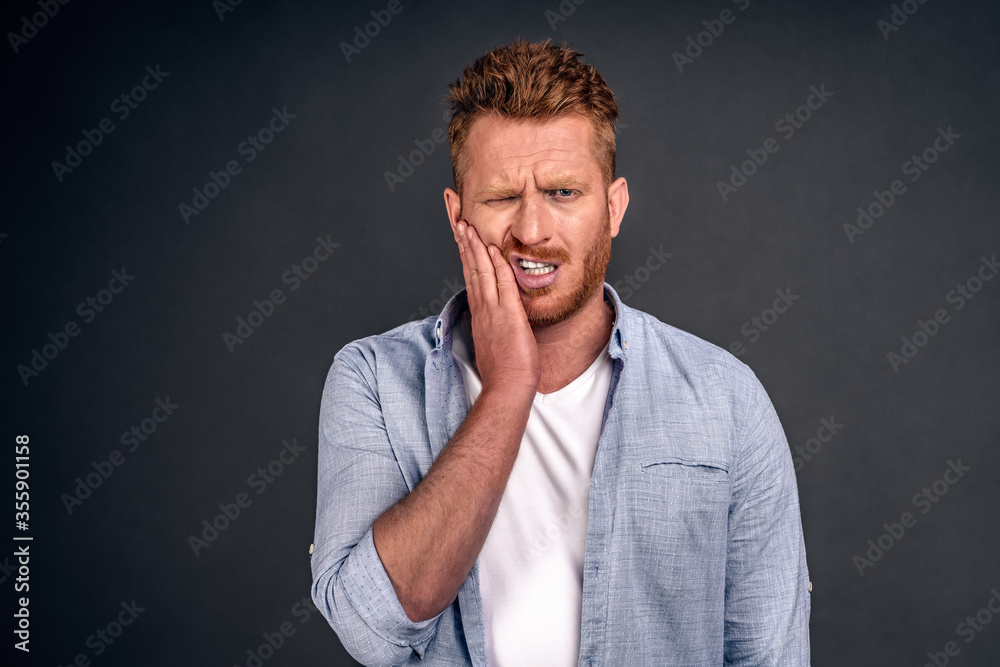 Photo of Caucasian redhaired man suffers painful toothache, has rotten tooth, needs to visit dentist, keeps eyes closed from pain, dressed in casual shirt, isolated over gray background, touches cheek