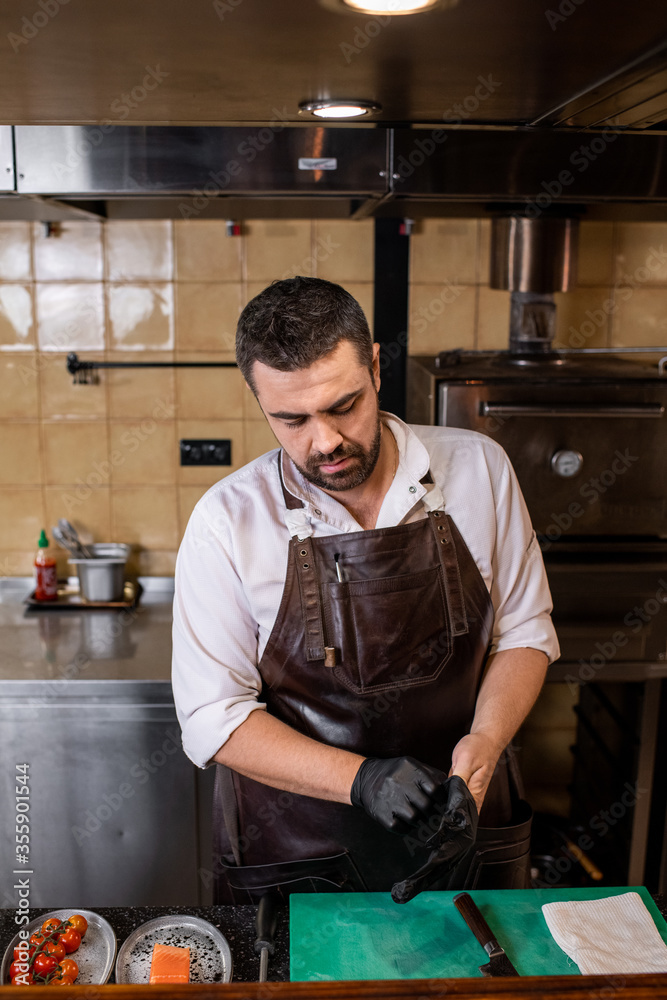 Busy bearded chef in leather apron standing at counter and putting gloves on hands before cooking Busy bearded chef in leather apron standing at counter and putting gloves on hands before cooking