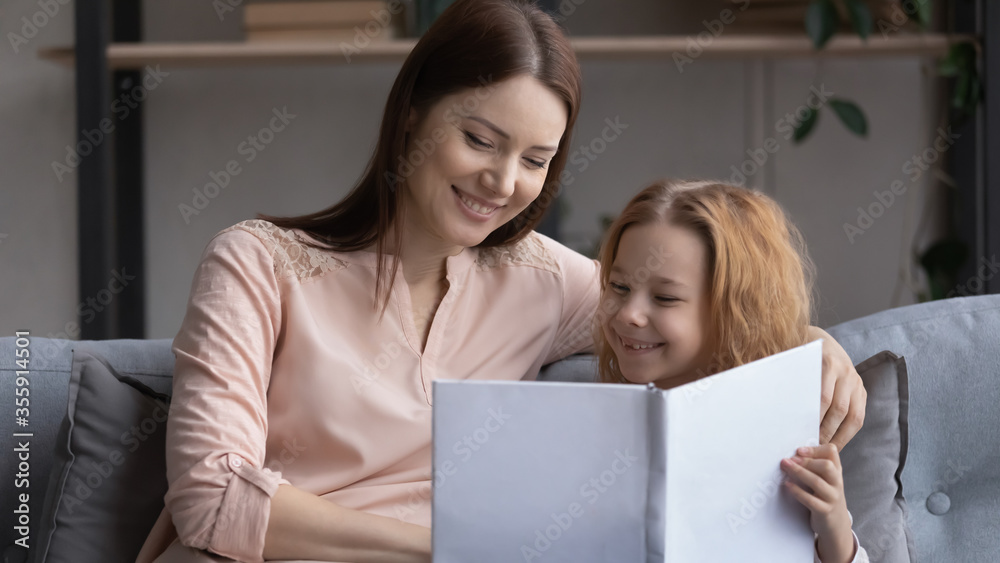 Smiling young Caucasian mom and little preschooler daughter sit on sofa at home reading book together, happy mother or nanny enjoy interesting fairytale story with small girl child, education concept Smiling young Caucasian mom and little preschooler daughter sit on sofa at home reading book together, happy mother or nanny enjoy interesting fairytale story with small girl child, education concept