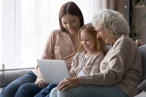 Smiling three generations of women relax on sofa in living room watch video movie on laptop, happy little girl with mom and senior grandmother rest on couch at home using modern computer together