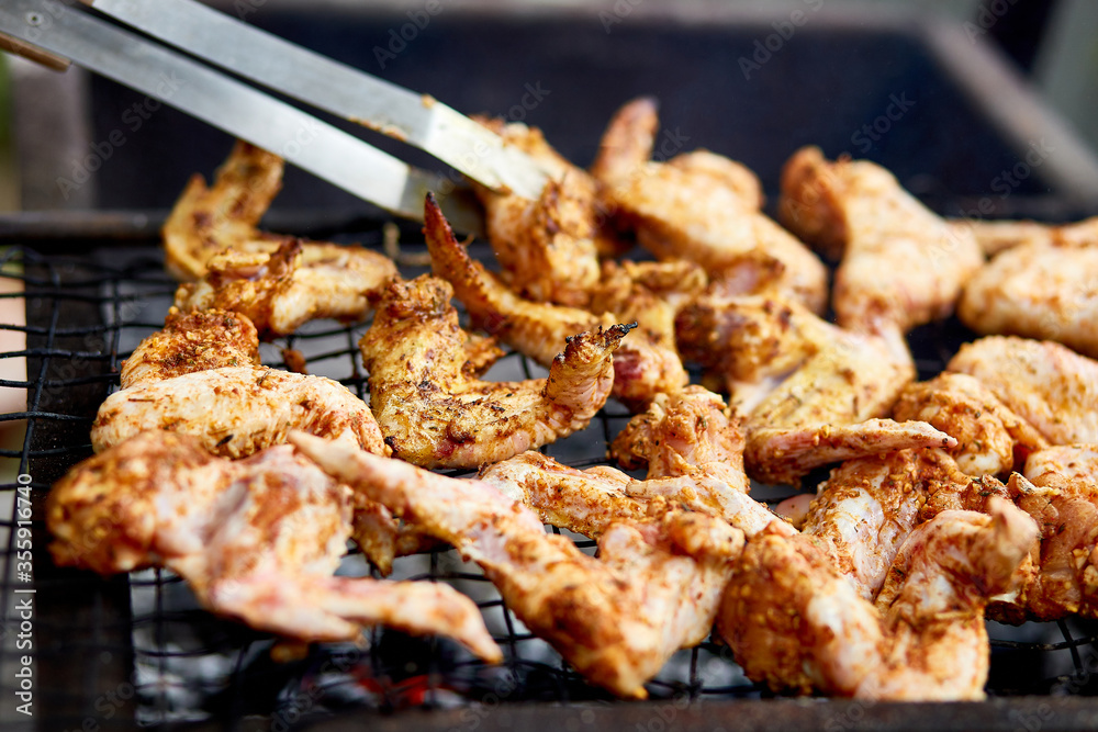 Man using metal tongs to turning chicken wings which are being grilled on barbecue, outdoor, meat is fried in mangal barbecue grill, Chef cooking in backyard at home.