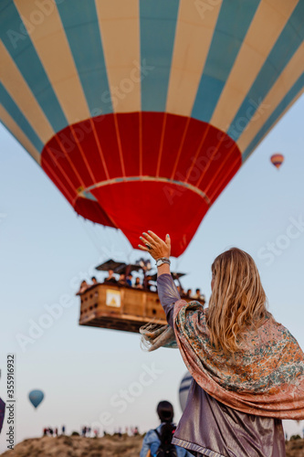 Beautiful blonde woman greets flying hot air balloon at dawn in Cappadocia