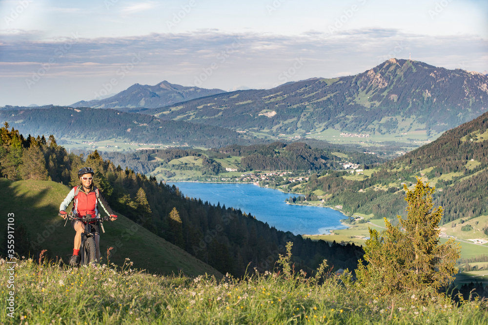 pretty senior woman riding her electric mountain bike in warm dawn sunlight heights of Salmas hight above Oberstaufen, with spectacular view on lake Alpsee,Allgau Alps, Bavaria Germany 
