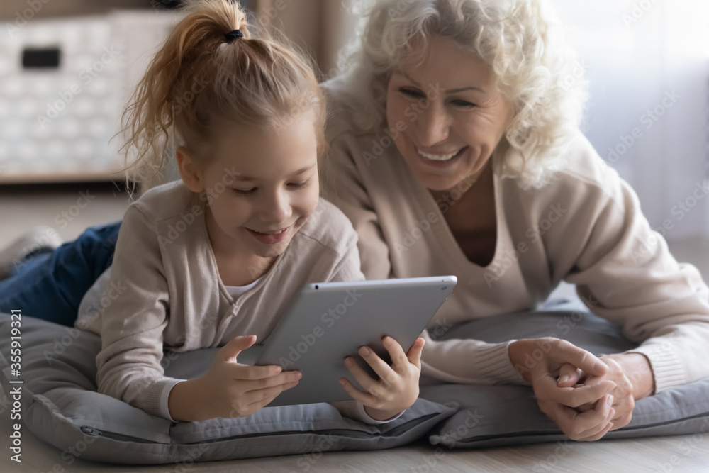 Playful happy elderly grandmother and little granddaughter relax lying on floor at home watching video pad gadget, smiling mature grandparent have fun play using modern tablet with small grandchild - obrazy, fototapety, plakaty 