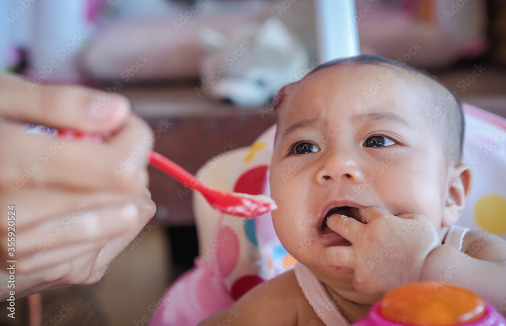 Sad cute baby refusing to eat baby food with a red spoon on a baby chair and crying while mother hand-feeding food at home. Dirty face of unhappy kid. Sad cute baby refusing to eat baby food with a red spoon on a baby chair and crying while mother hand-feeding food at home. Dirty face of unhappy kid.