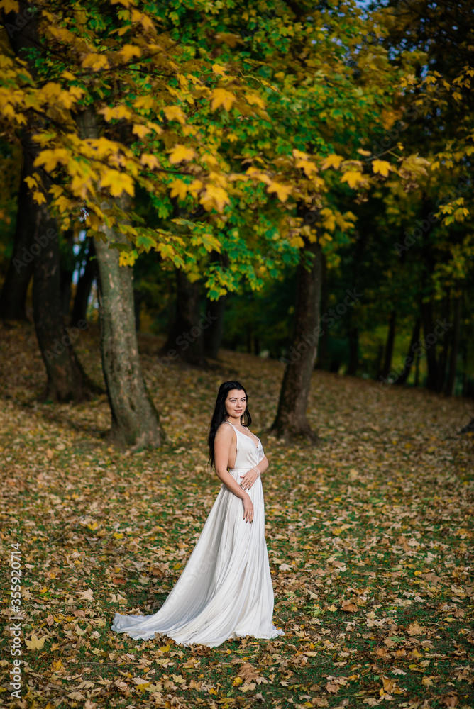 Young beautiful brunette female in a long white dress in autumn park.
