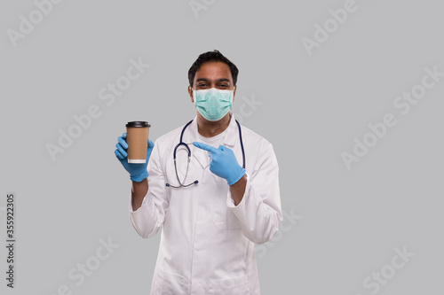 Indian Man Doctor Pointing at Coffee Take Away Cup Wearing Medical Mask and Gloves Isolated. Indian Doctor Holding Coffee To Go Cup.