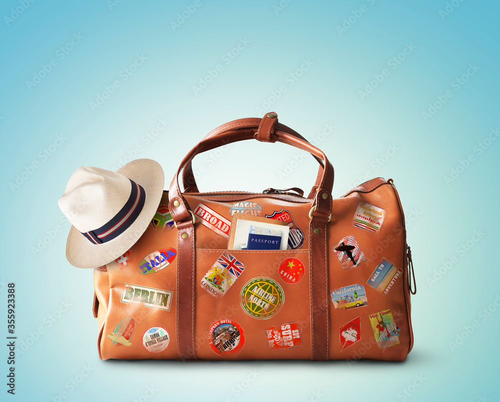 Vacation concept, large classic brown leather travel bag with hat - obrazy, fototapety, plakaty 
