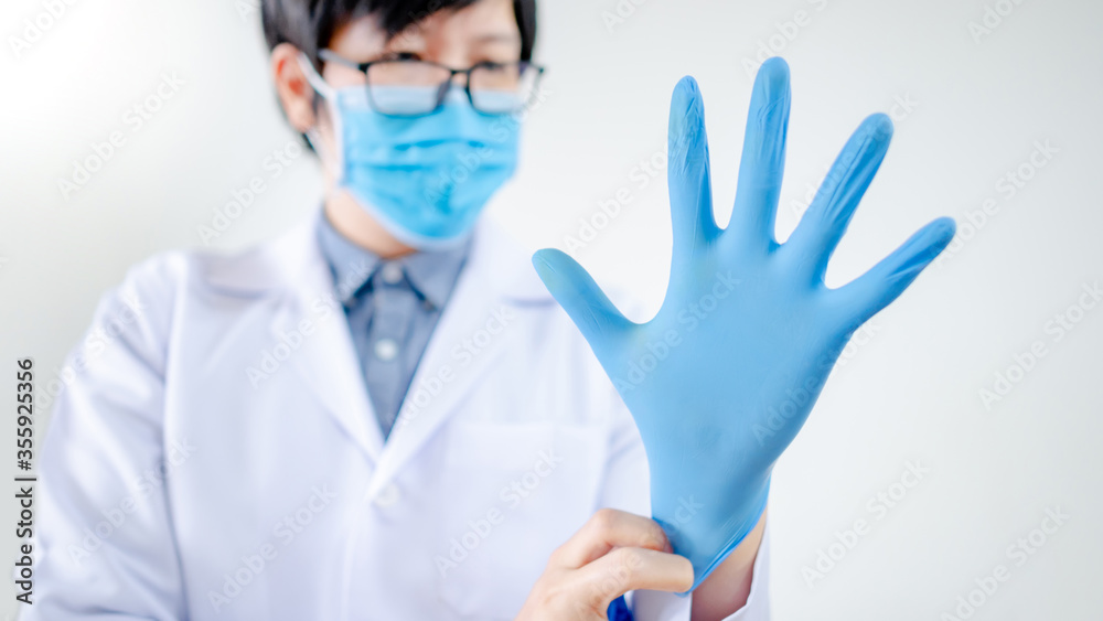 Male Asian doctor or Physician man in white uniform and blue surgical mask putting disposable latex gloves on hand while working in hospital clinic. Coronavirus (COVID-19) infection prevention. Male Asian doctor or Physician man in white uniform and blue surgical mask putting disposable latex gloves on hand while working in hospital clinic. Coronavirus (COVID-19) infection prevention.