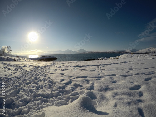 long snowy field with footprints with beautiful bay, blue fjord and sky on a sunny winter day in the arctic circle