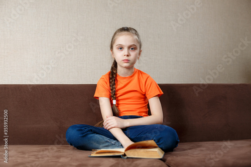 Cute Little Girl Reading a Book on Sofa in lotus Position at Home. Distance Learning Education. Digital detox.