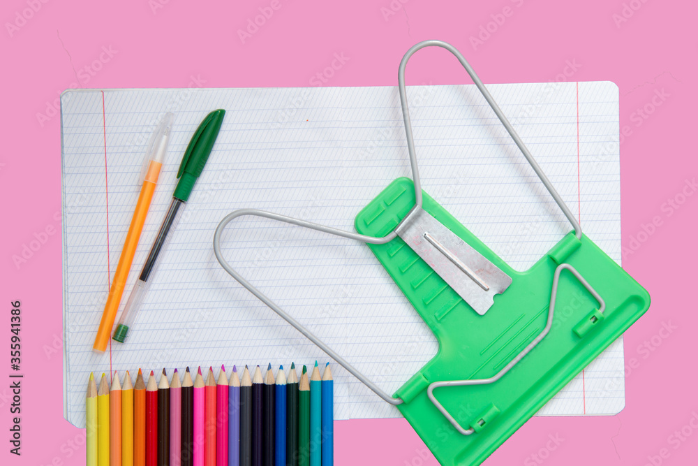 On a pink background is an open notebook in thin and oblique stripes, as well as pens, pencils and a book stand. School background. On a pink background is an open notebook in thin and oblique stripes, as well as pens, pencils and a book stand. School background.