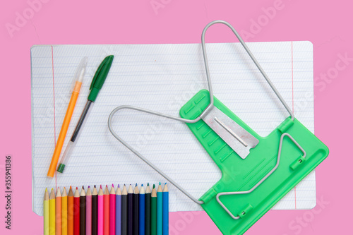 On a pink background is an open notebook in thin and oblique stripes, as well as pens, pencils and a book stand. School background.