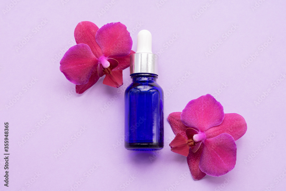 Blue glass bottle filled by essence or serum with orchid extract on purple background with bright blossoming orchids phalaenopsis. Eco cosmetic concept Blue glass bottle filled by essence or serum with orchid extract on purple background with bright blossoming orchids phalaenopsis. Eco cosmetic concept